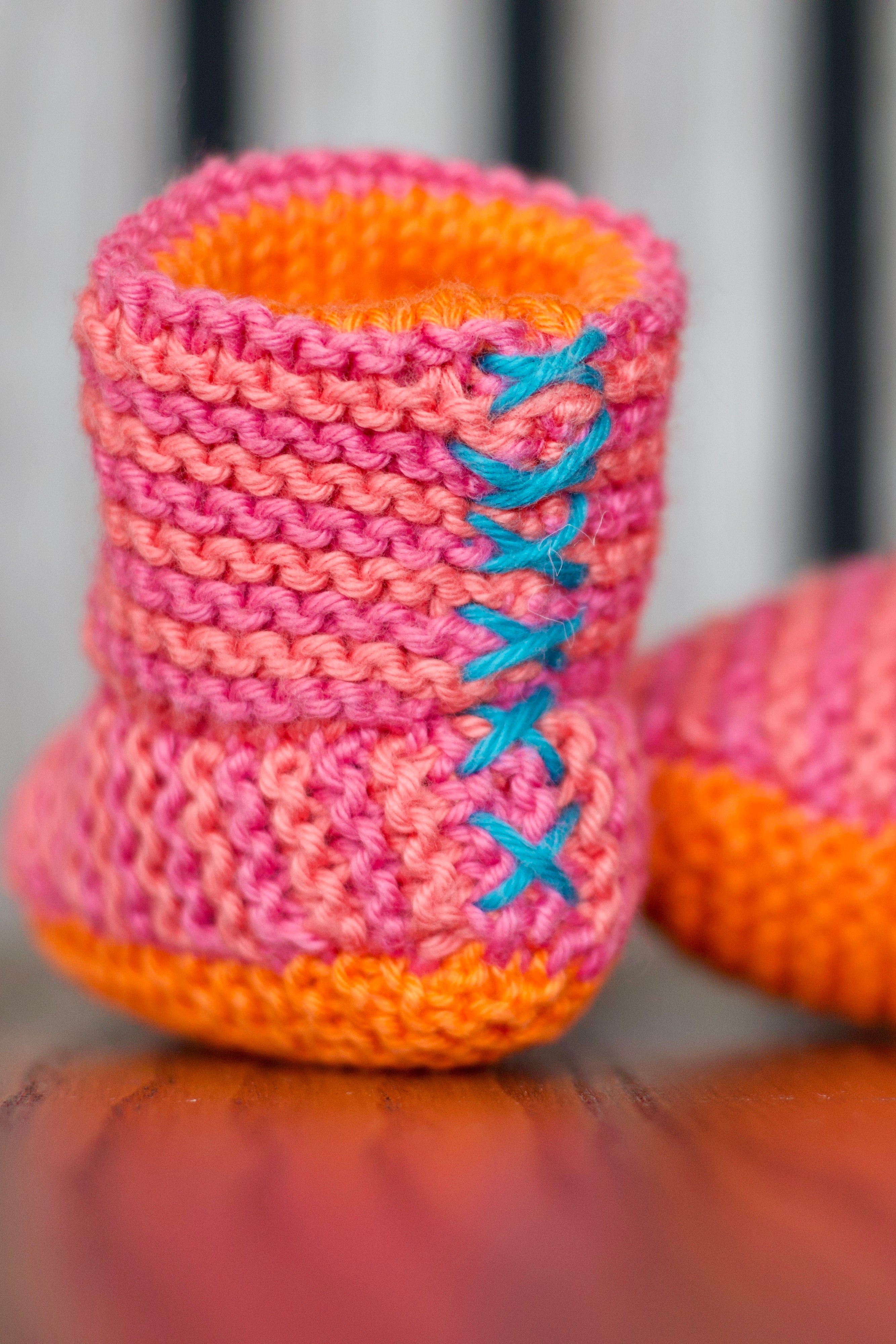 A close-up of a hand-knitted baby bootie in pink and orange with blue embroidered laces, sitting on a wooden surface with another bootie blurred in the background.