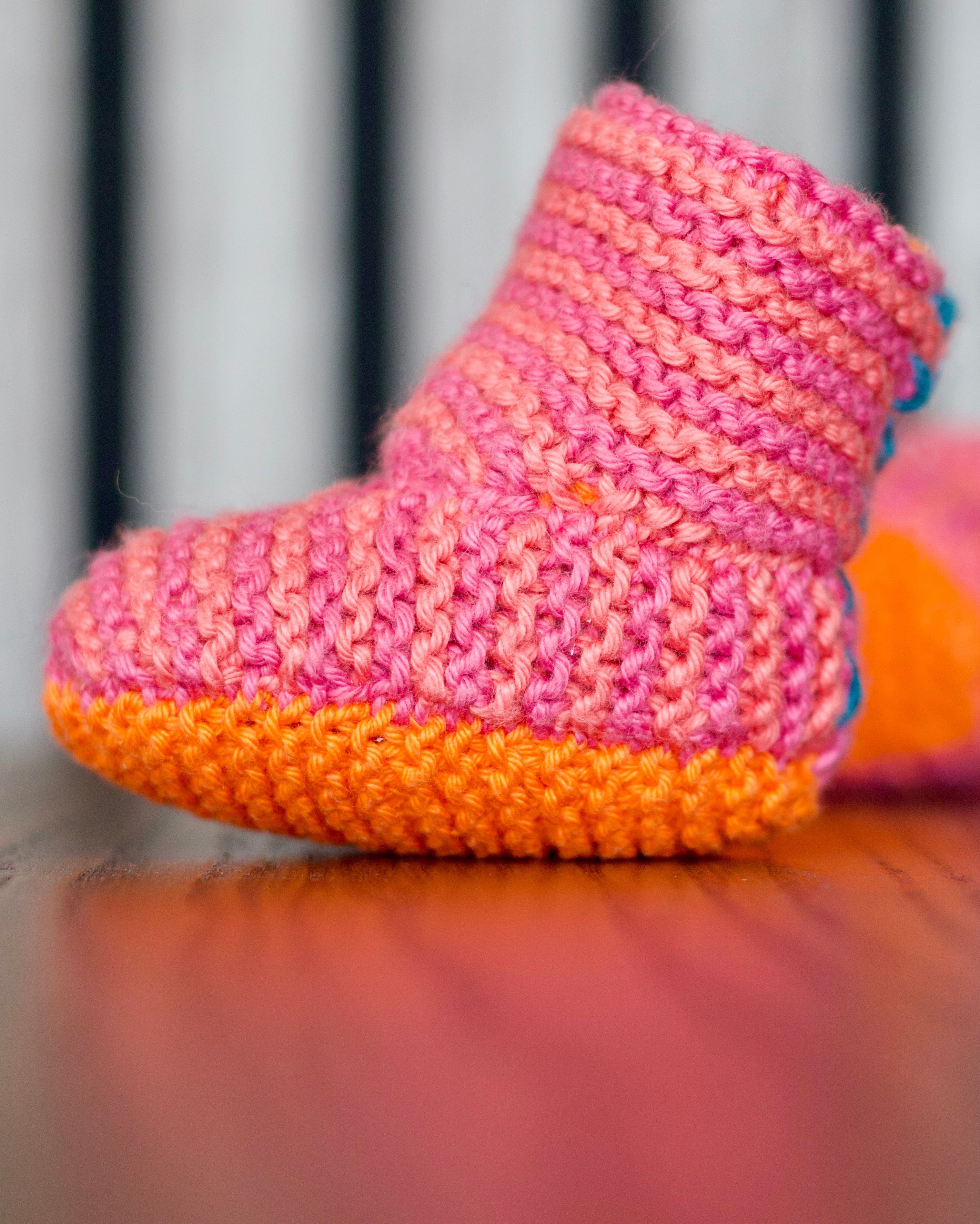 A close-up of a hand-knitted baby bootie in pink and orange, resting on a wooden surface with a blurred background.