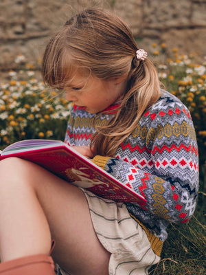 A young girl sitting on the ground reading a book while wearing The Faraway Gang's 'Daydreamer' Knitted Jumper.