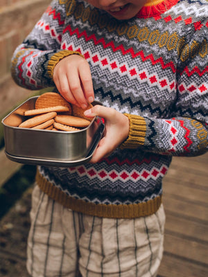A young girl wearing 'The Daydreamer' Knitted Jumper from The Faraway Gang with a tin of biscuits.