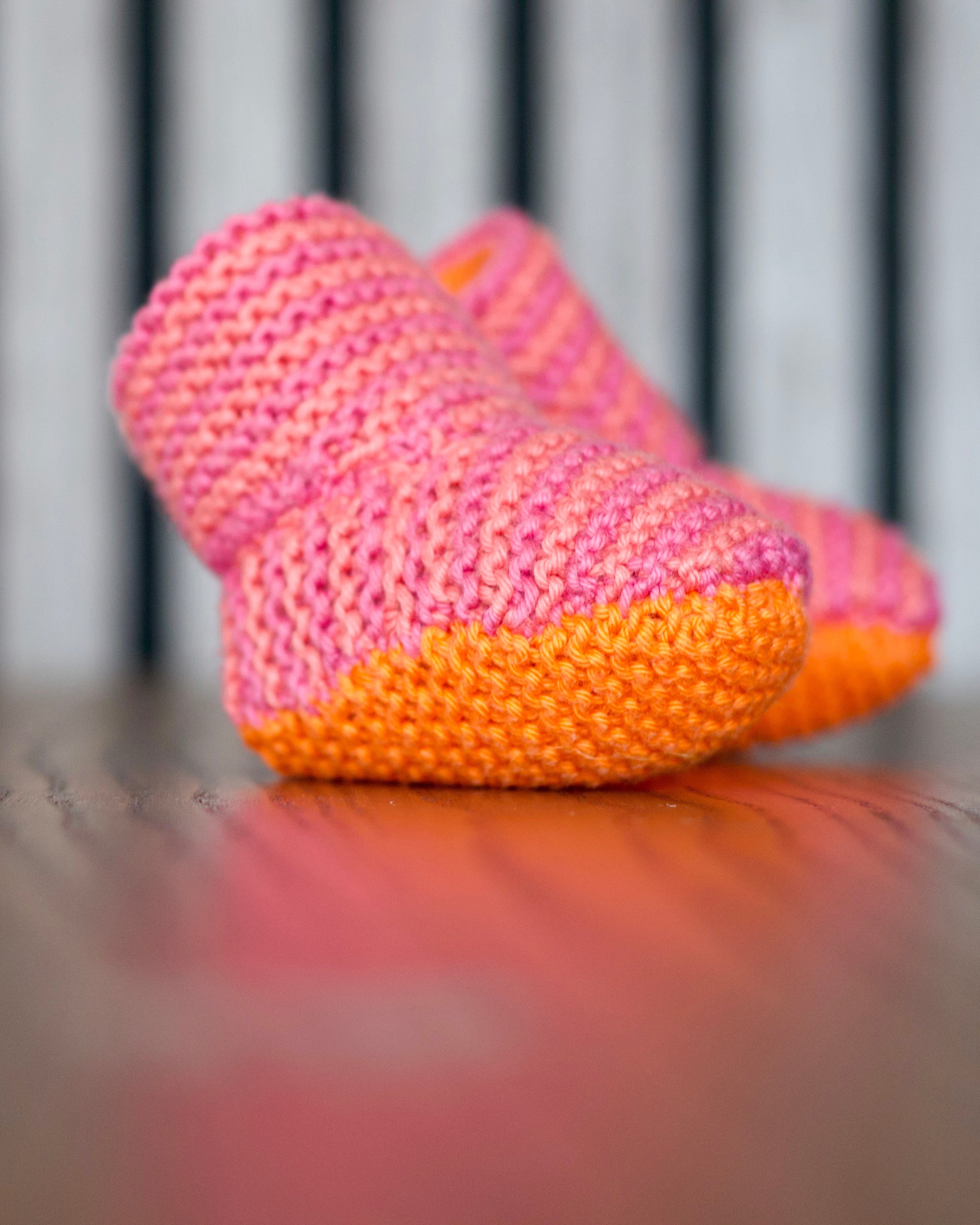 A pair of hand-knit baby booties in pink and orange, resting on a wooden surface with a blurred background.