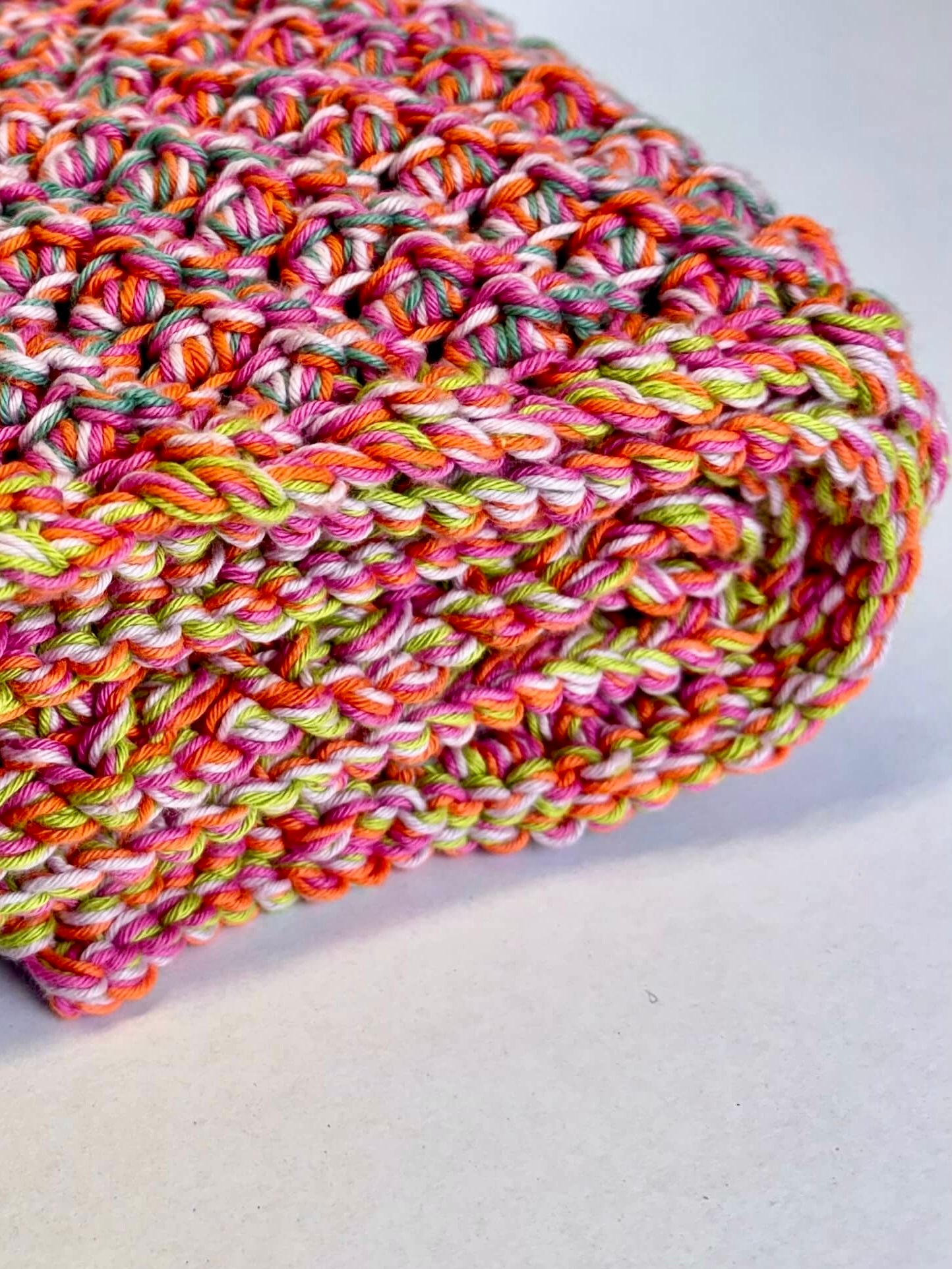 A close-up of a chunky knit blanket or throw made from variegated yarn in bright pink, orange, yellow, green, and white, folded on a light gray surface.