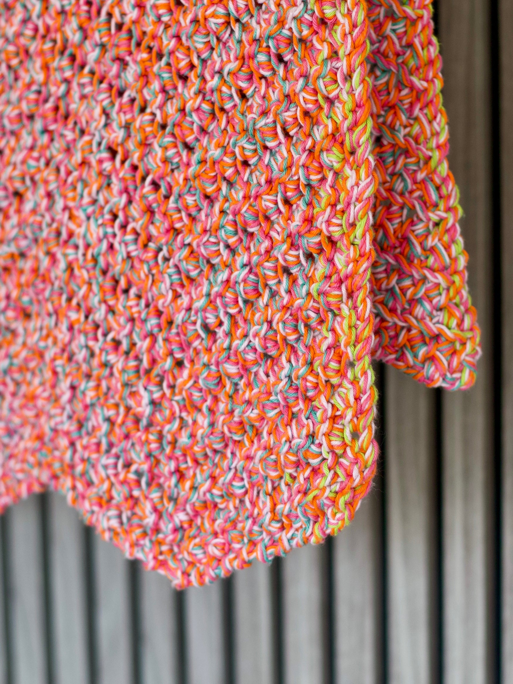 A close-up of a chunky knit blanket or throw in a variegated multicolor yarn, featuring orange, pink, yellow, and blue threads, draped against a gray wooden slatted background.