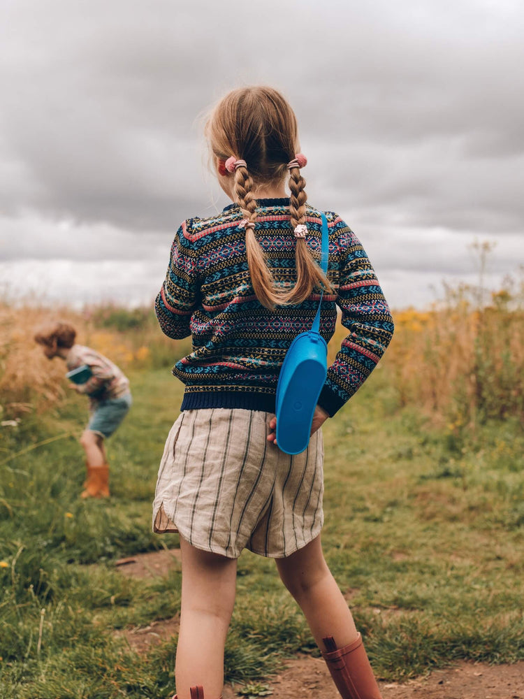 A young girl is stood in a field and is wearing 'Explorer' Knitted Jumper by The Faraway Gang and facing away from us.