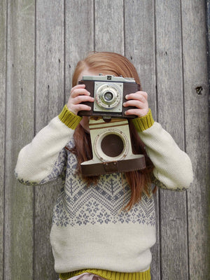 A young girl holding a camera up to her face, The Faraway Gang's 'Storyteller' Knitted Jumper.