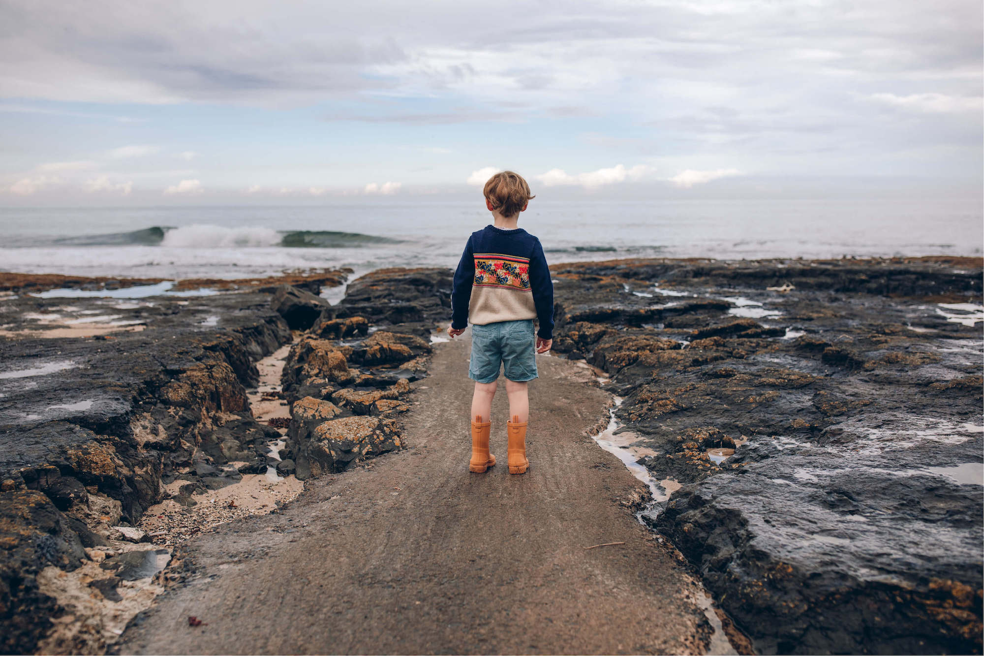 a young boy standing on a rocky beach next to the ocean.