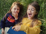 Two girls wearing The Faraway Gang printed sweatshirts, laughing and rolling in the grass.