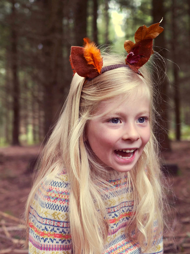 A young girl is sitting in the woods wearing 'Explorer' Knitted Jumper by The Faraway Gang and laughing.