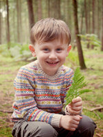 A young boy sitting in the woods wearing 'Explorer' Knitted Jumper by The Faraway Gang and holding a leaf.
