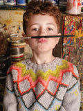 Child holding a paintbrush to their face with a colorful sweater in an art studio.