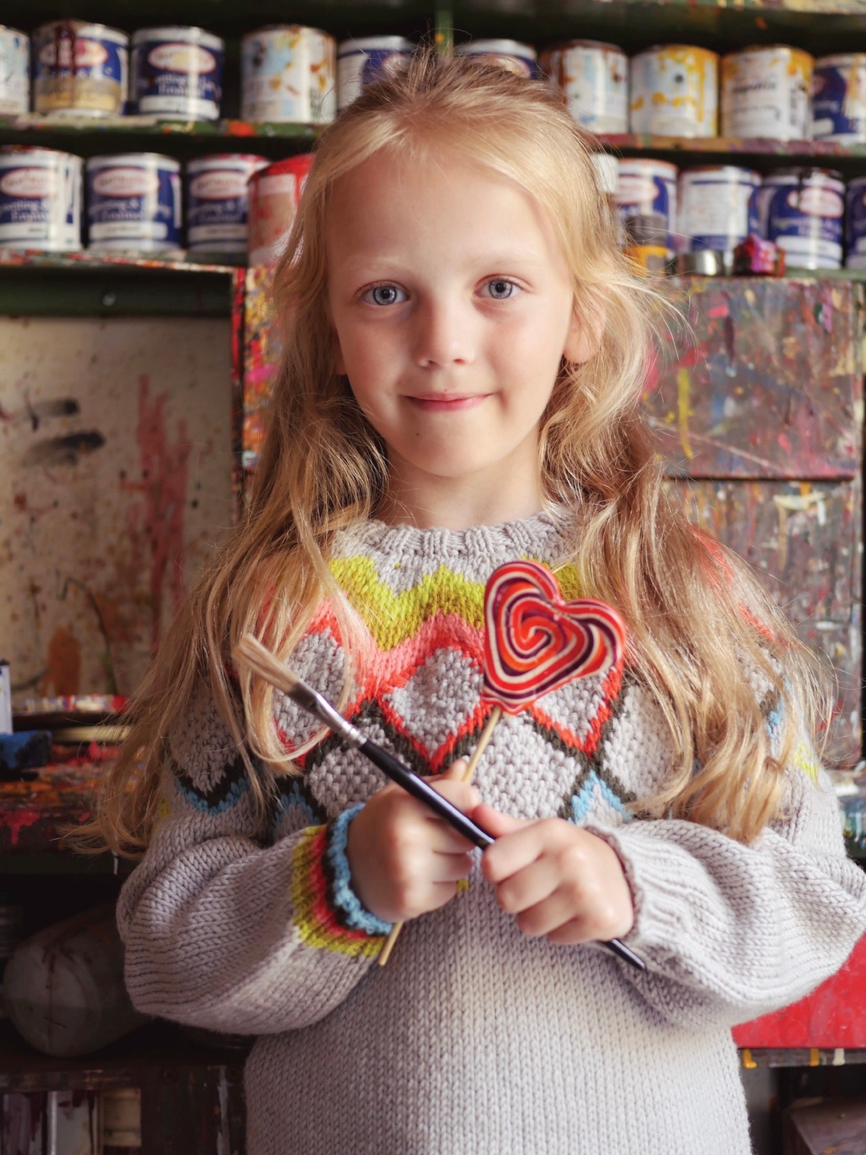 Young girl wearing a colorful sweater with a heart design, holding a paintbrush in an art studio.
