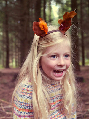 A young girl is sitting in the woods wearing 'Explorer' Knitted Jumper by The Faraway Gang and laughing.