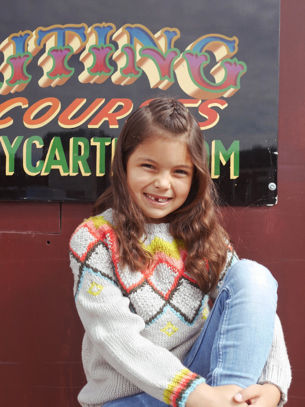 Young girl in a colorful sweater sitting in front of a colorful sign.