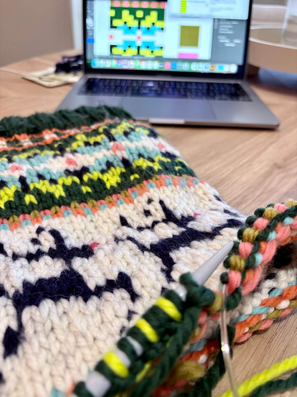 Knitted multicolored fabric with knitting needles on a wooden table next to a laptop displaying a pattern.