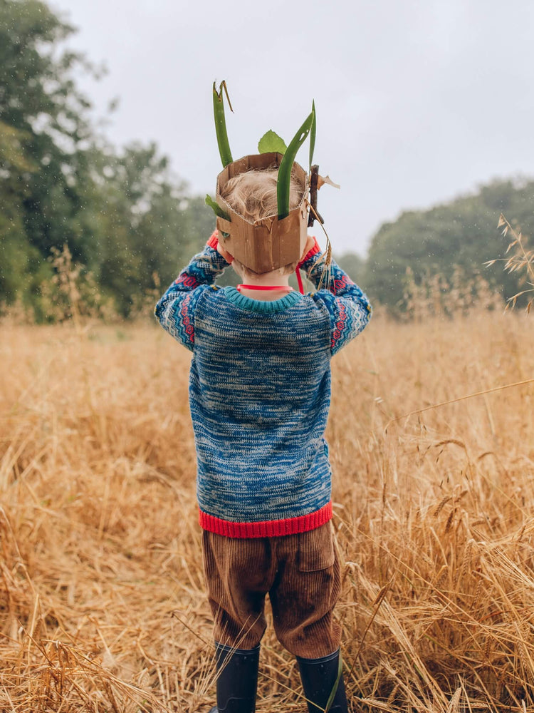 A little boy standing in a field wearing 'The Daydreamer' Knitted Jumper by The Faraway Gang with a cardboard crown on his head.