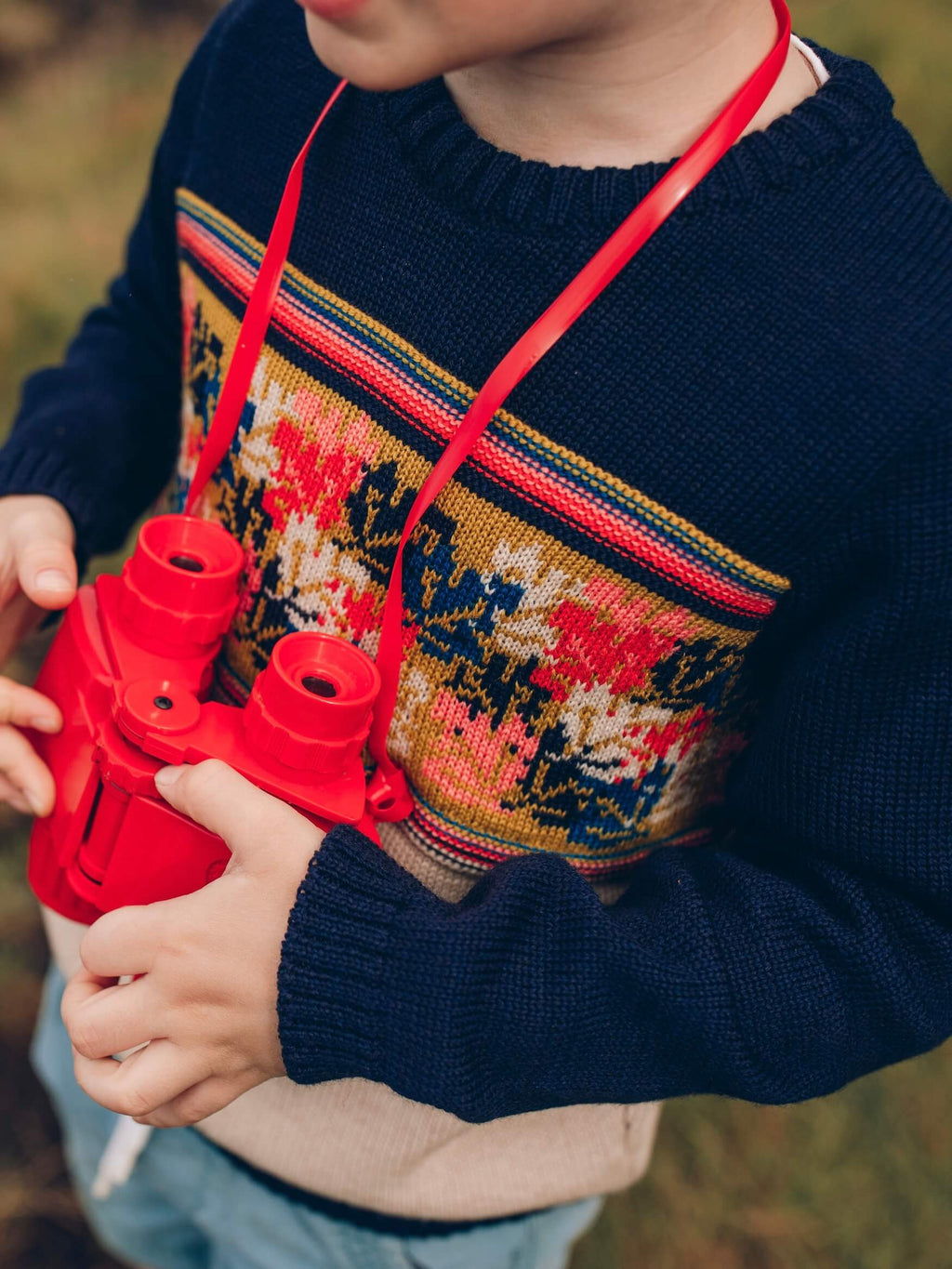 A young boy holding a pair of red binoculars, wearing The Faraway Gang's The 'Adventurer' Knitted Jumper.
