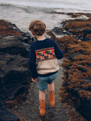 A little boy wearing The 'Adventurer' Knitted Jumper by The Faraway Gang standing on a rocky beach next to the ocean.