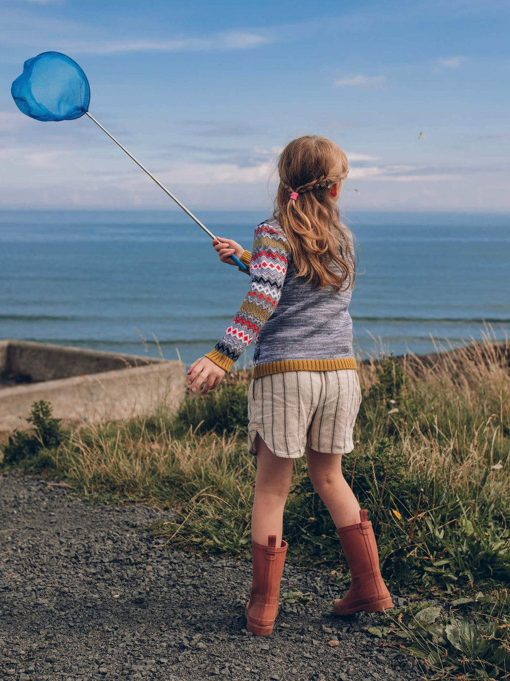A little girl wearing 'The Daydreamer' Knitted Jumper by The Faraway Gang, holding a blue fishing net near the ocean.