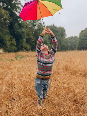 A young boy wearing 'The Daydreamer' Knitted Jumper by The Faraway Gang and holding an umbrella above their head in a field.