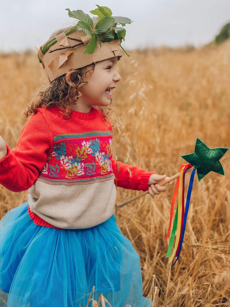 A little girl standing in a field wearing The Faraway Gang's 'Adventurer' Knitted Jumper in colour pomegranate with a homemade cardboard crown on her head and carrying a wand.