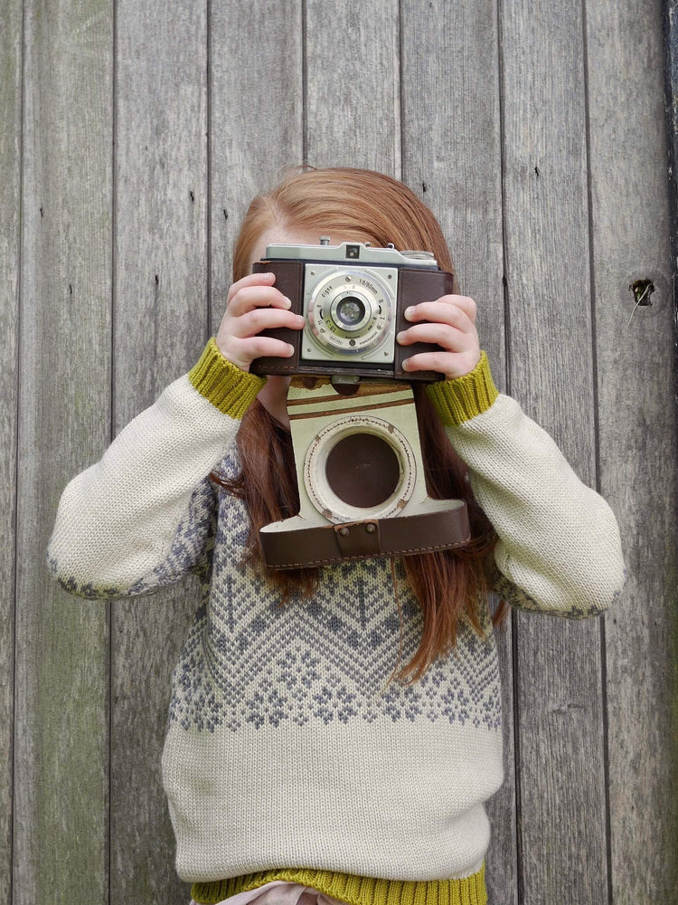 A young girl holding a camera up to her face, The Faraway Gang's 'Storyteller' Knitted Jumper.
