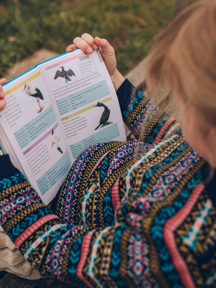A young girl is wearing 'Explorer' Knitted Jumper by The Faraway Gang and reading a bird book.