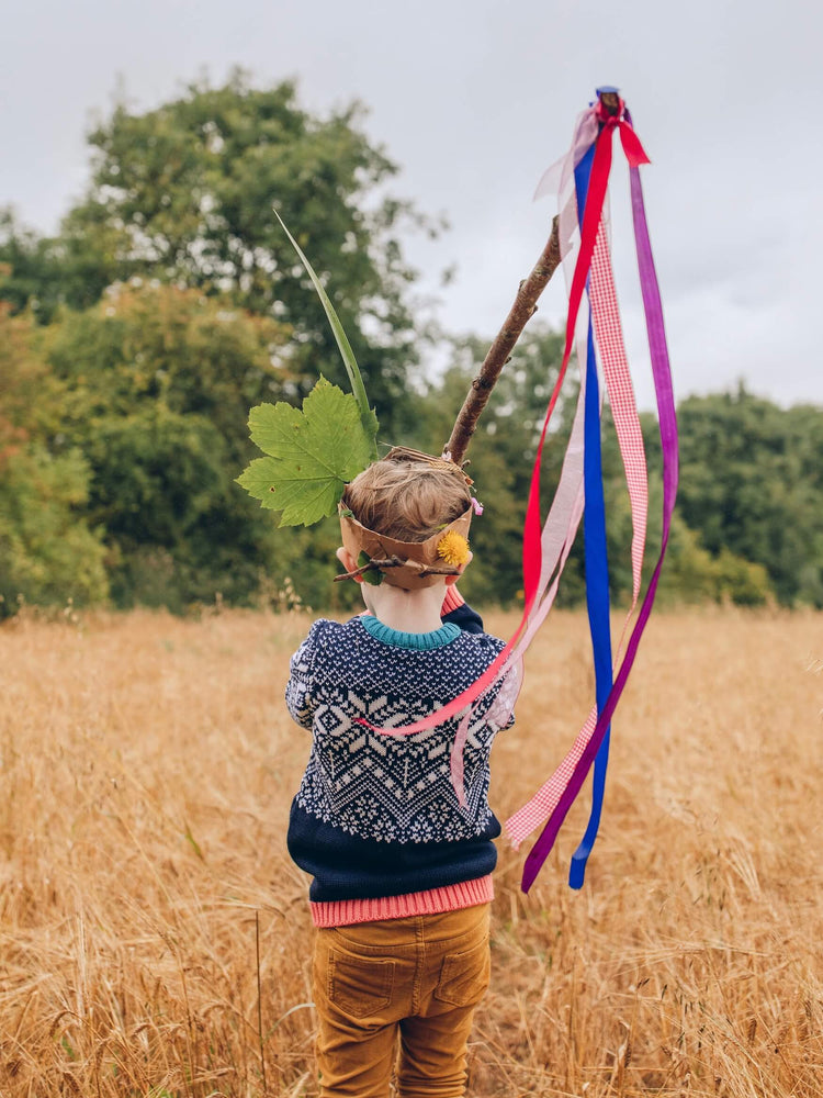 A child standing in a field wearing The Faraway Gang's 'Storyteller' Jumper holding a stick with colourful ribbons hanging from it.