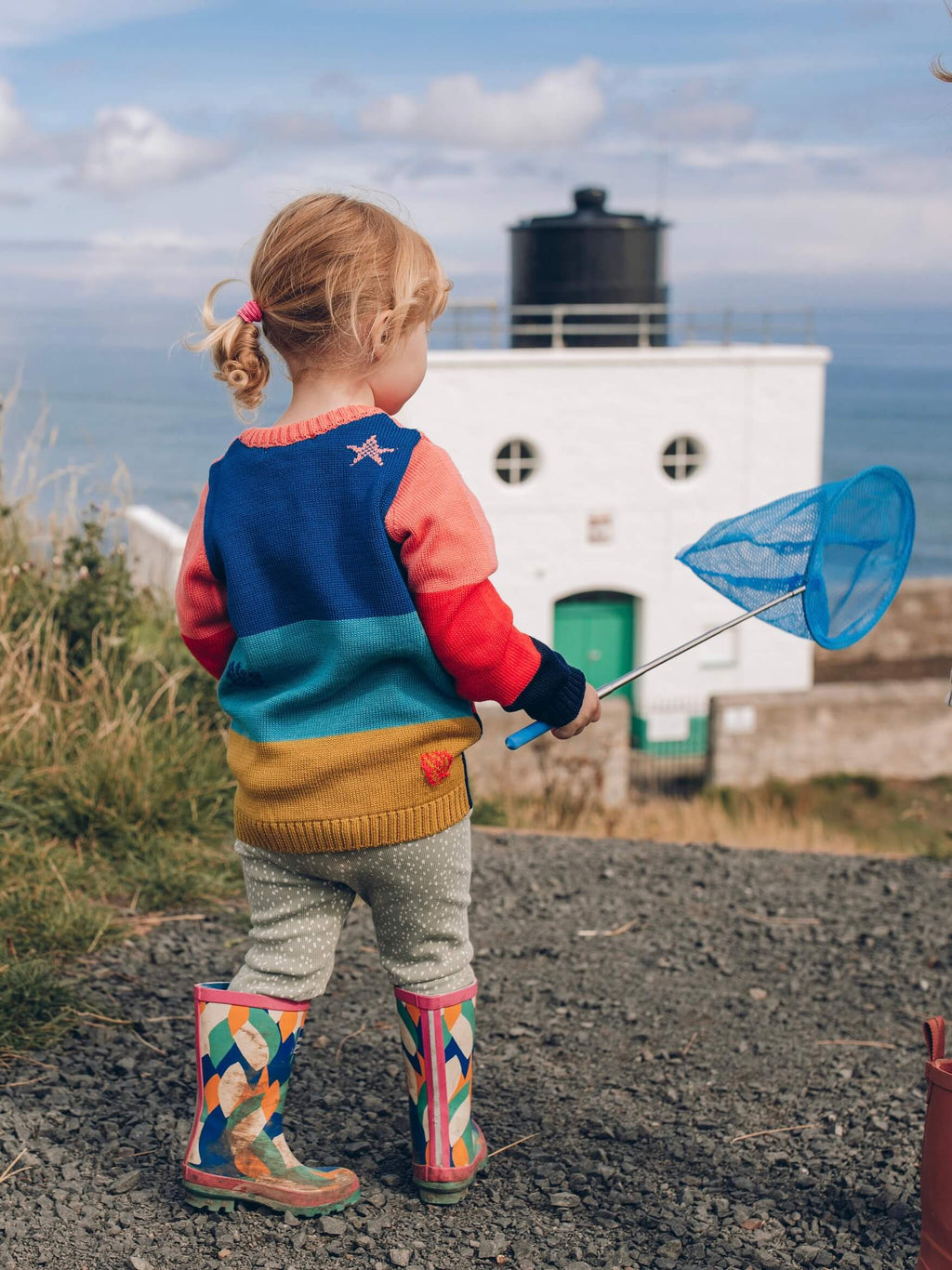 A little girl holding a blue fishing net near a lighthouse, dressed in 'The Stargazer' Knitted Jumper from The Faraway Gang.
