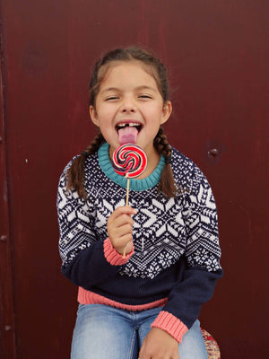 A little girl standing in the woods looking up wearing 'The Storyteller' Knitted Jumper by The Faraway Gang licking a lolly pop..