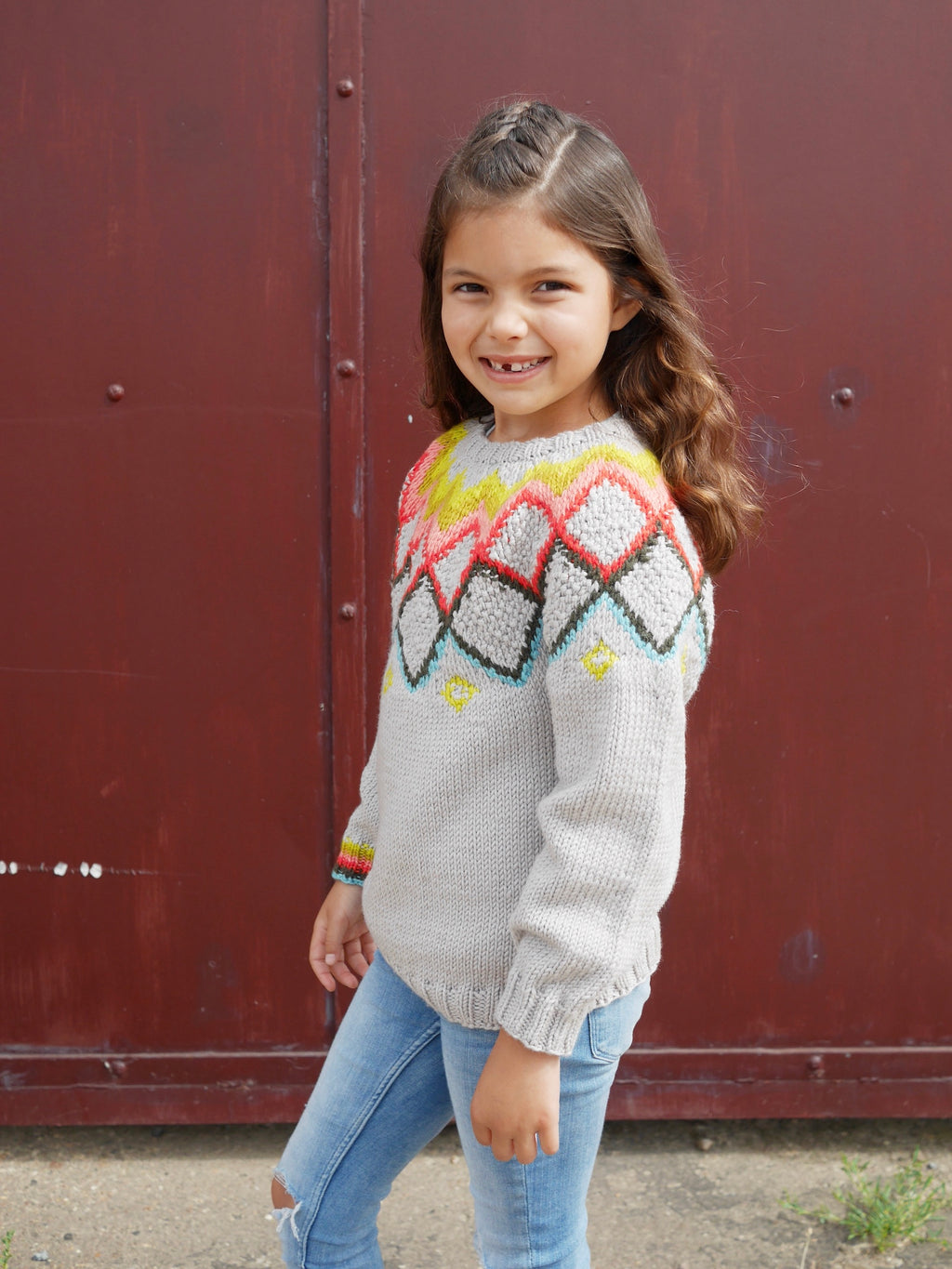 Young girl wearing a colorful sweater standing in front of a red wall.